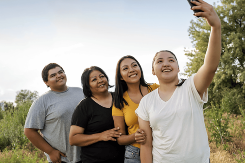A family of four standing closely together outdoors smiling and taking a group selfie.