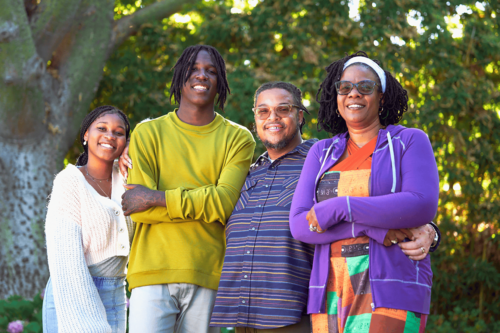 A family of two adults and two teens standing close together and smiling in front of a tree.