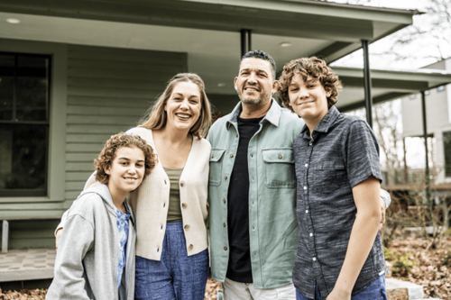 A family of two adults and two children standing close together and smiling in front of a house.