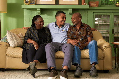 A teen smiling and sitting between two parents on a couch who are smiling and looking at each other.