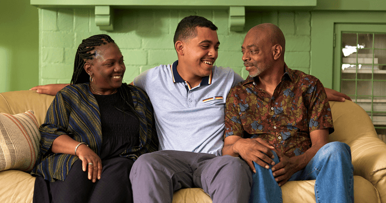 A teen smiling and sitting between two parents on a couch who are smiling and looking at each other.